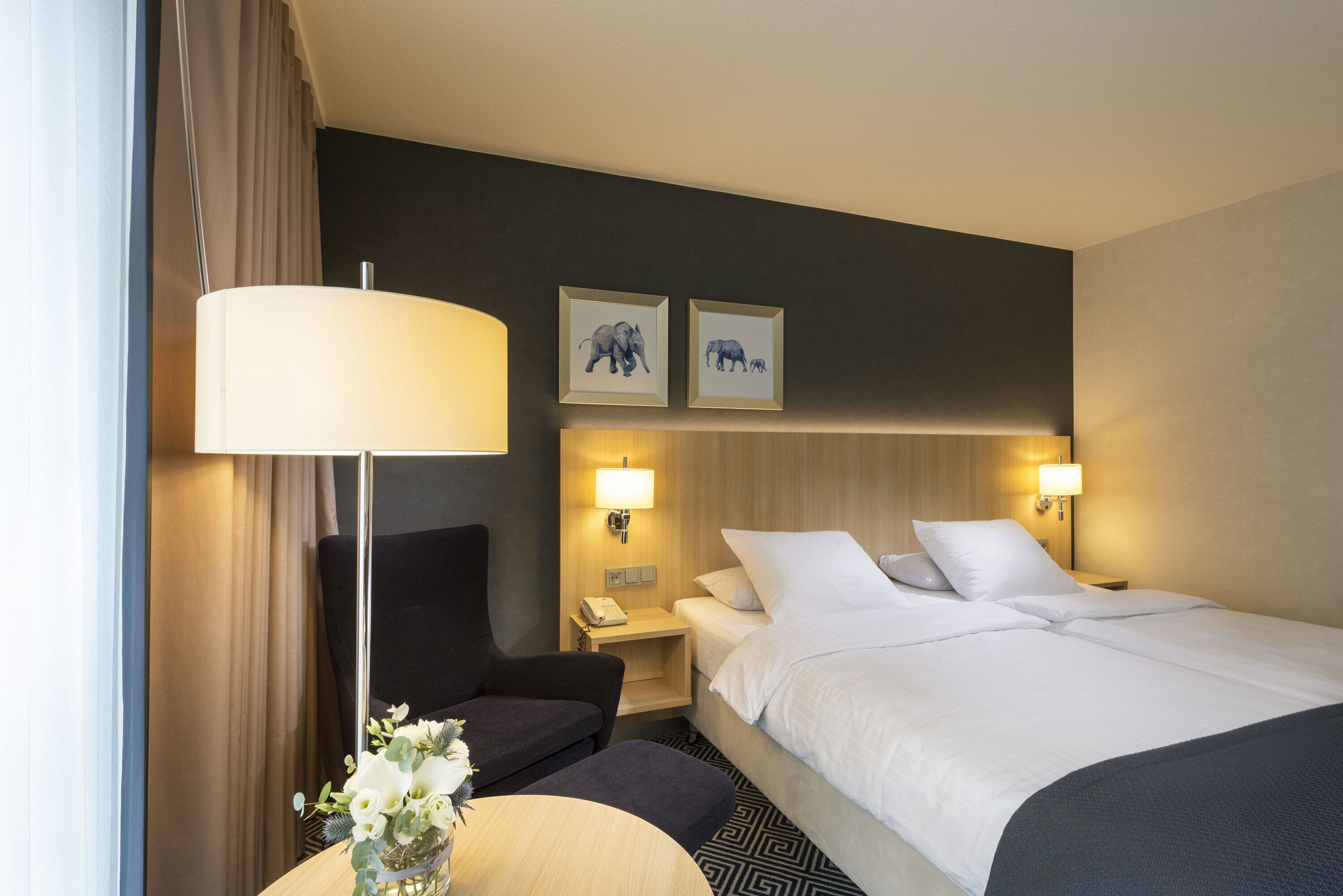 A hotel room with a bed, two bedside tables, a desk with a chair, a lamp, and two framed pictures on the wall.
