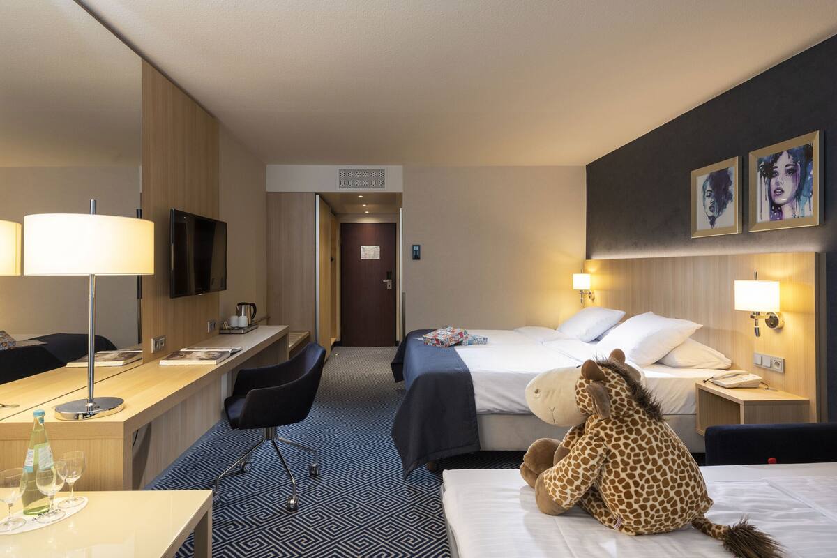 A hotel room with a bed, desk, chair, and a giraffe plush toy.
