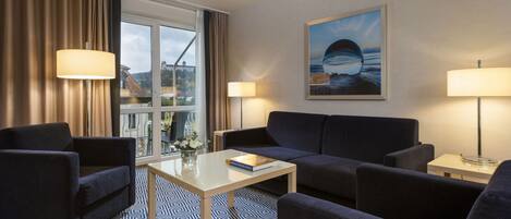 A modern hotel room with a sofa, two armchairs, a coffee table, and a painting of a coastal scene.