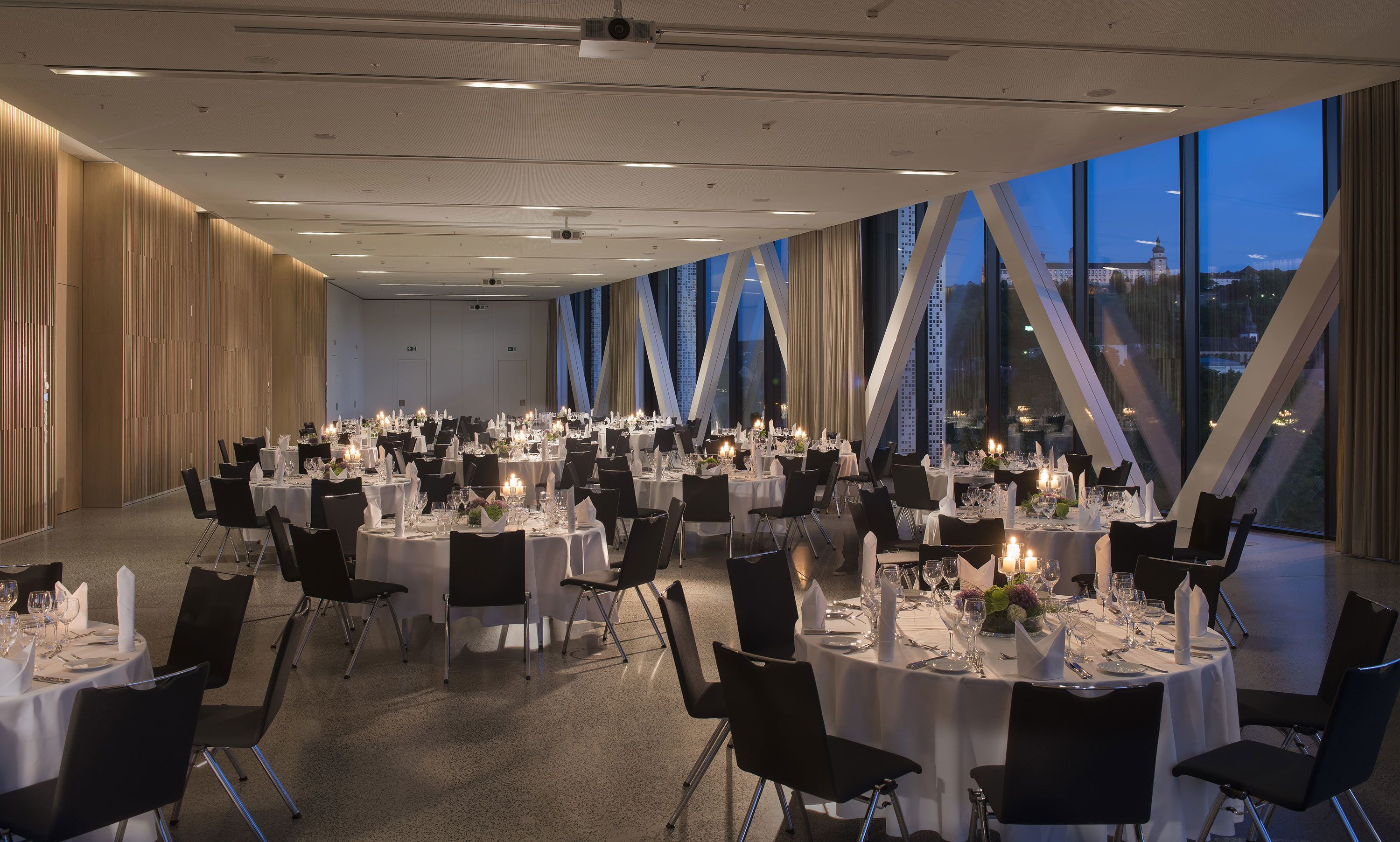 A spacious banquet hall with round tables set for dining, large windows offering a city view, and a modern ceiling design.