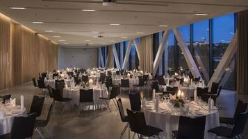 A spacious banquet hall with round tables set for dining, large windows offering a city view, and a modern ceiling design.