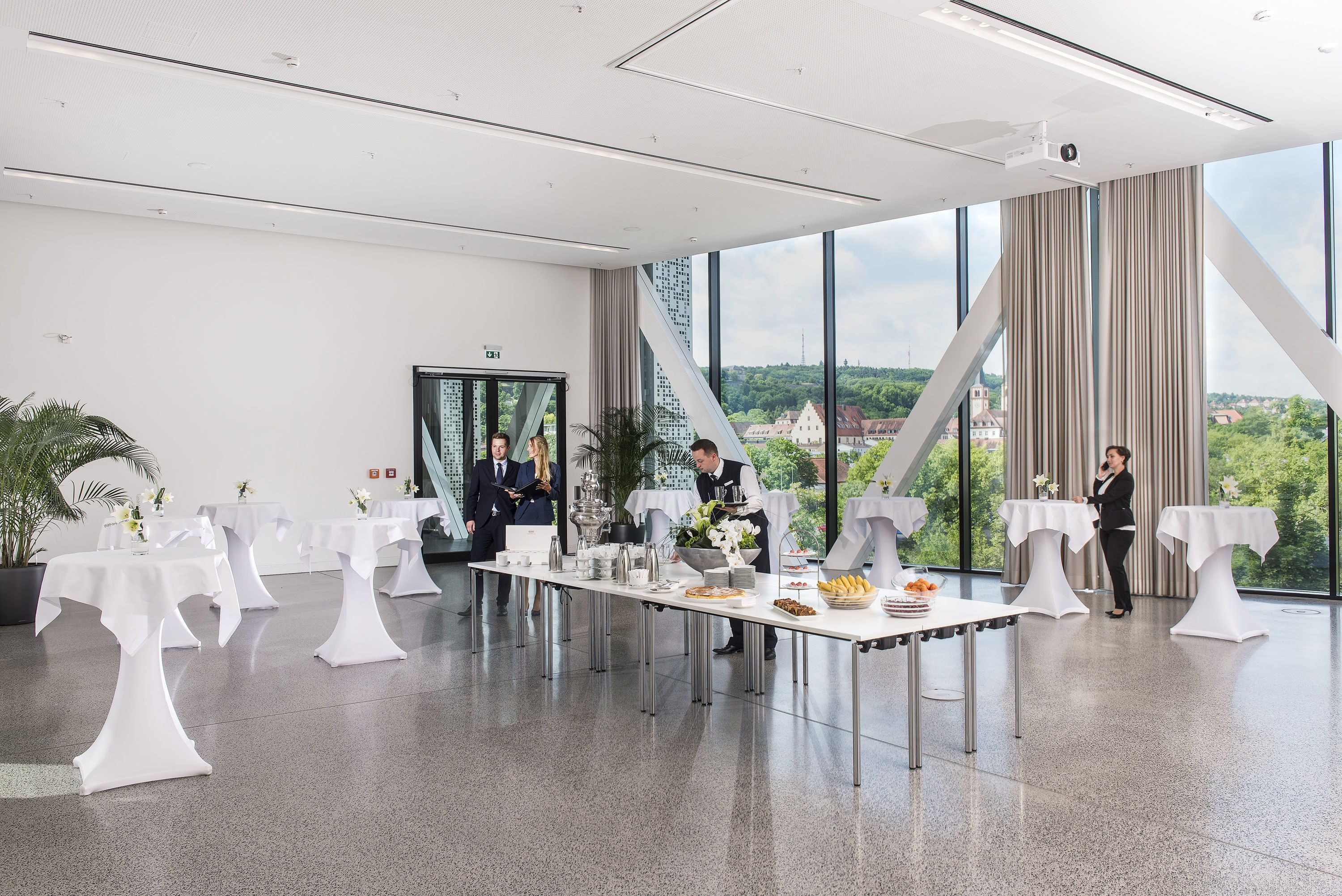 Un salón de conferencias moderno con mesas blancas, un ventanal con vista a la ciudad y un proyector en el techo.