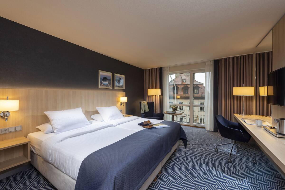 A hotel room with a large bed, a desk with a chair, a small table with a lamp, and a view of the city through the window.