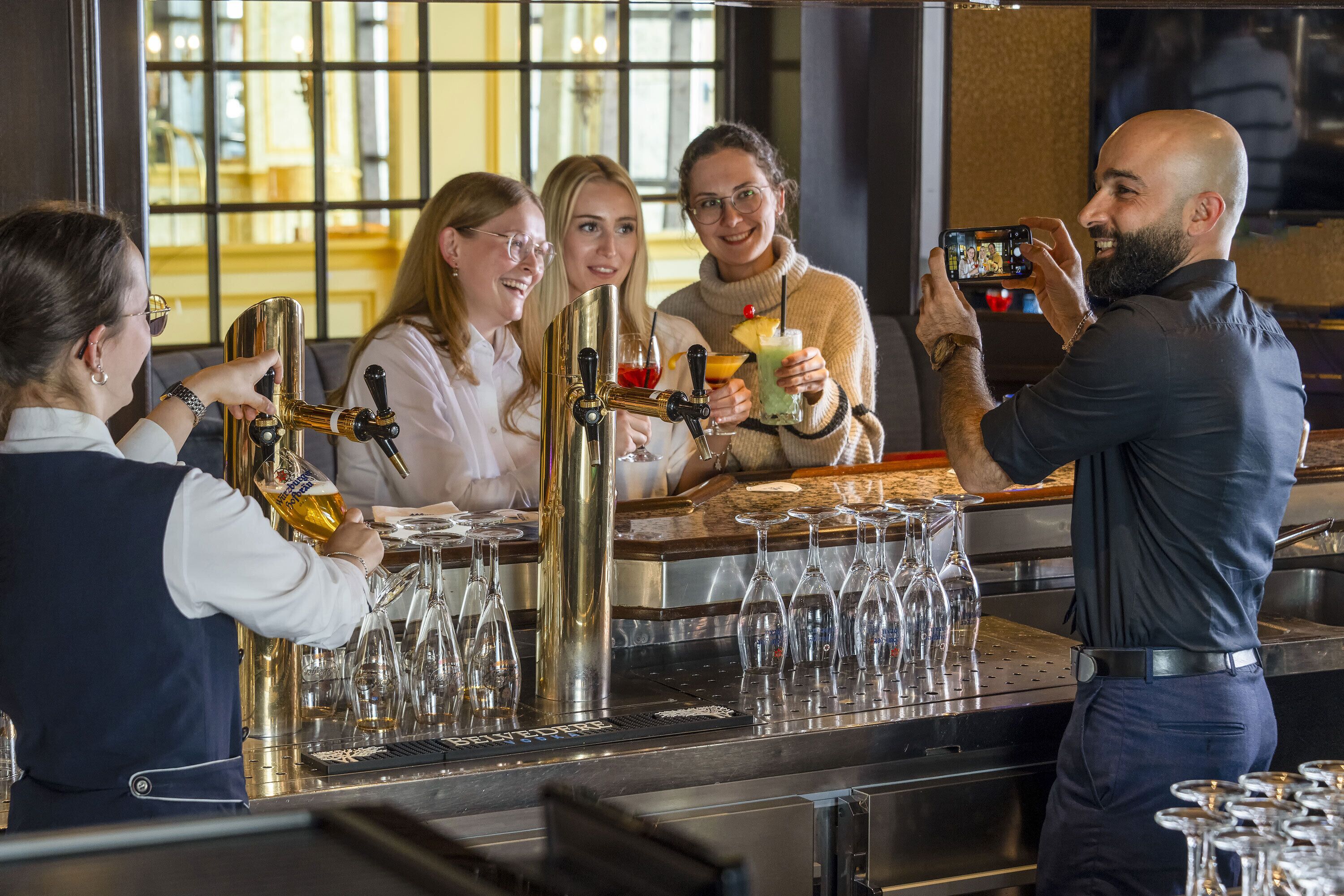 Una zona de bar con un barman preparando bebidas, un grupo de personas y alguien tomando una foto.