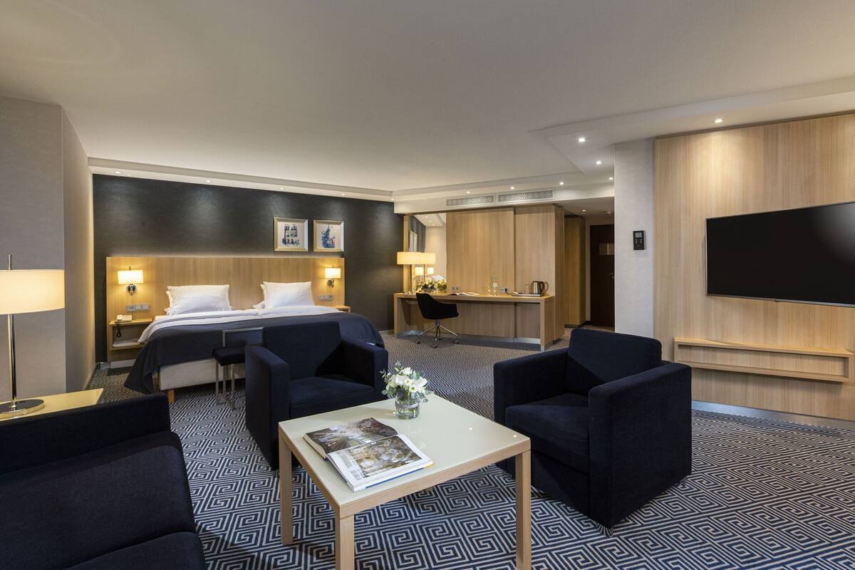 Ein Hotelzimmer mit einem Bett, zwei Sesseln, einem kleinen Tisch mit einer Zeitschrift, einem Schreibtisch mit Stuhl, einem an der Wand befestigten Fernseher und einem gemusterten Teppich.
