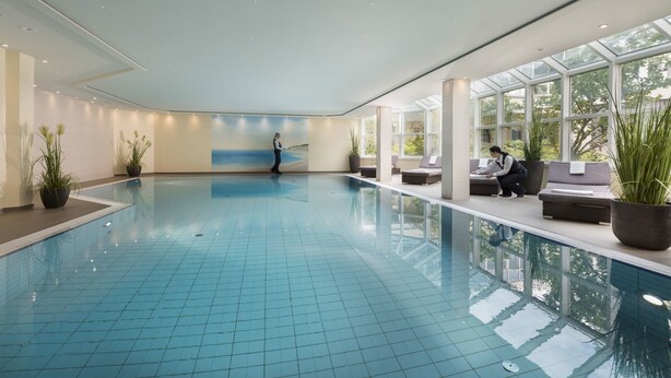 A large indoor swimming pool with a tiled floor, a person walking, and seating areas with plants. - Maritim Hotel Würzburg (Würzburg)