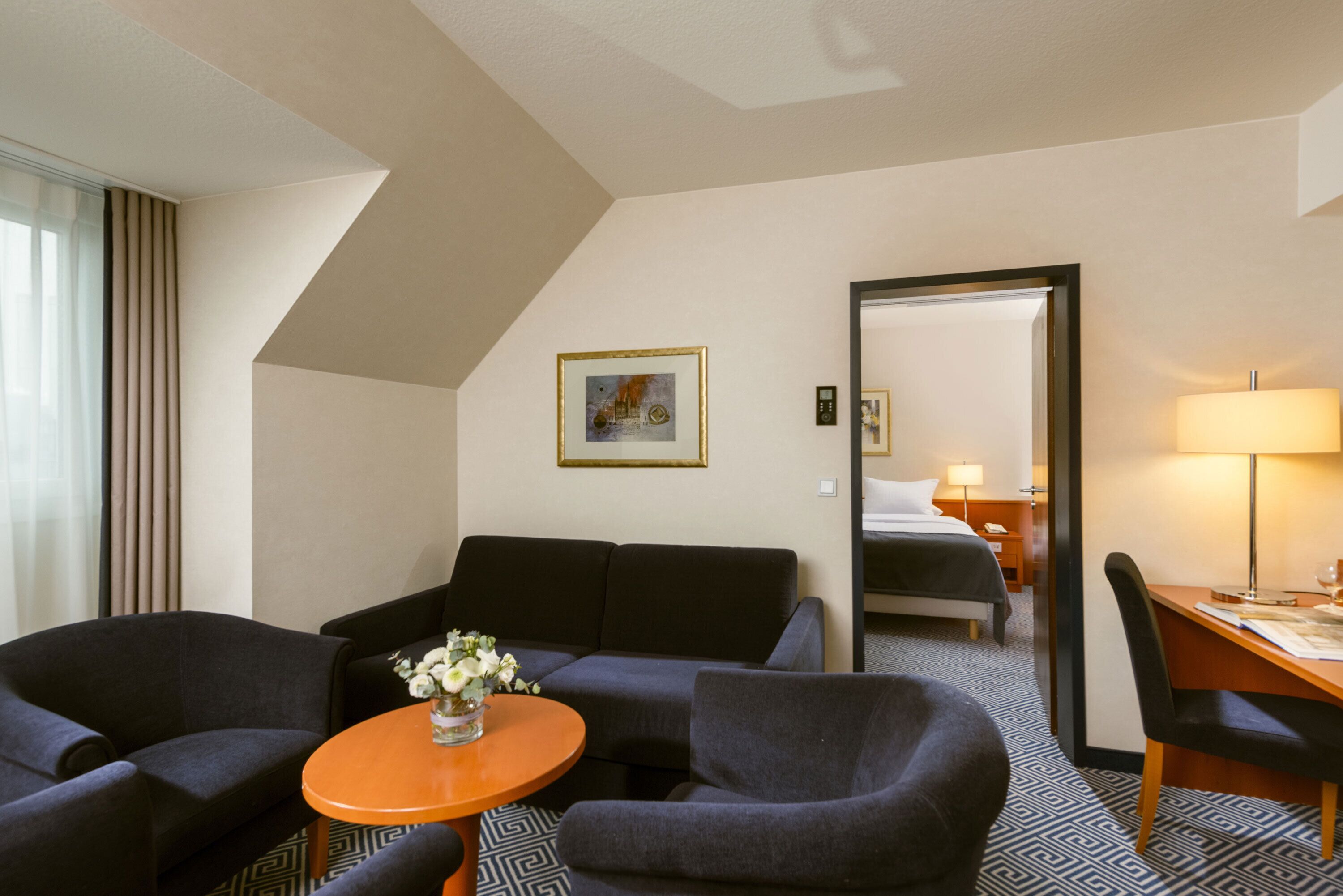 Habitación de hotel con sofá, sillones, una mesita con un jarrón de flores, cama, escritorio y lámpara.