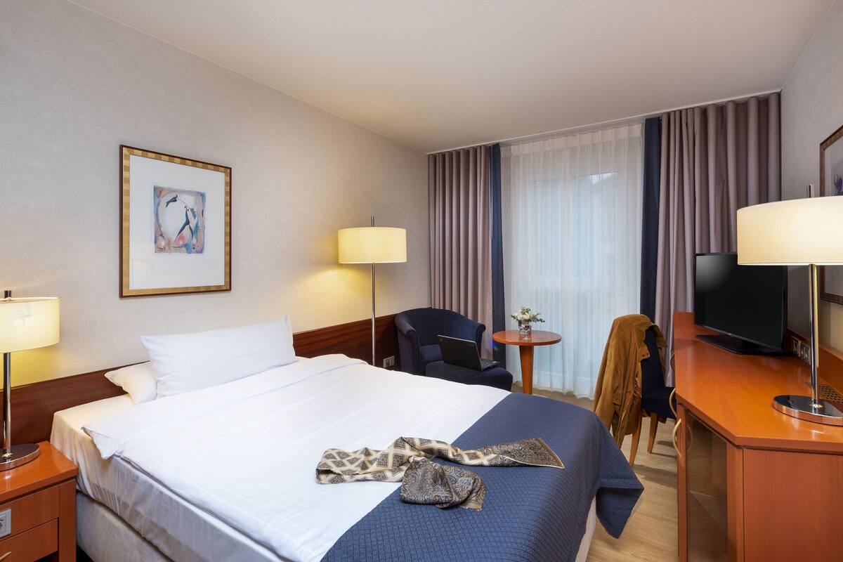 Ein Hotelzimmer mit einem großen Bett, zwei Nachttischen mit Lampen, einem Schreibtisch mit Stuhl, einem Fernseher und einem Fenster mit Vorhängen.