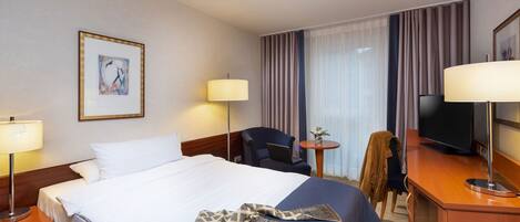 Habitación de hotel con una cama grande, dos mesitas de noche con lámparas, un escritorio con silla, un televisor y una ventana con cortinas.