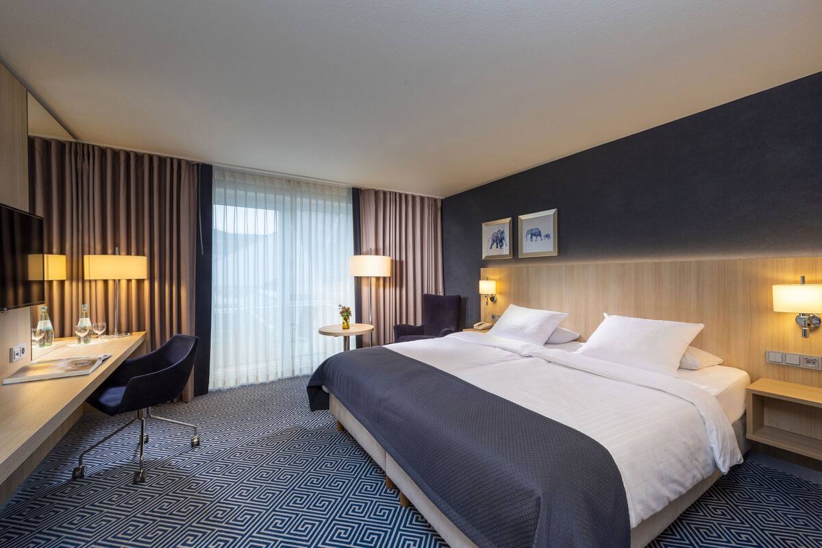 Ein Hotelzimmer mit einem großen Bett, einem Schreibtisch mit Stuhl, einem kleinen Tisch mit Lampe und einem großen Fenster mit Vorhängen.