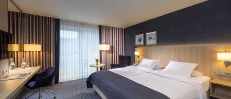 A hotel room with a large bed, a desk with a chair, a small table with a lamp, and a large window with curtains.