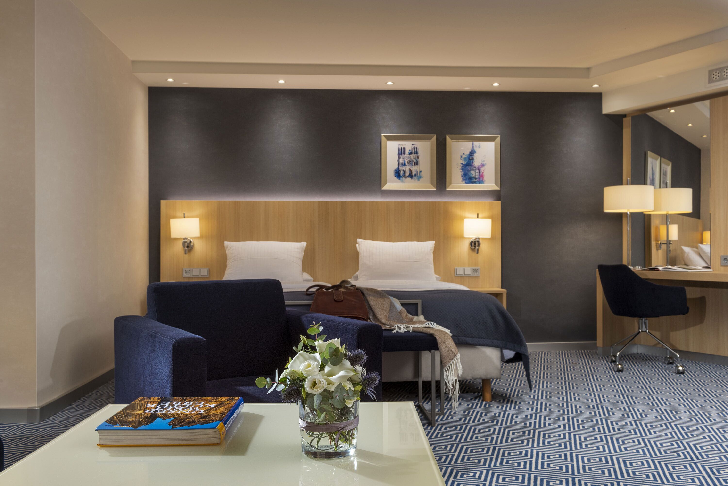 Una habitación de hotel con una cama, dos sillas, un escritorio y una mesa de centro. Hay un libro y un jarrón con flores sobre la mesa.
