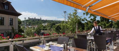 An outdoor café with tables and chairs, a waiter, and a scenic view of buildings and greenery in the background.