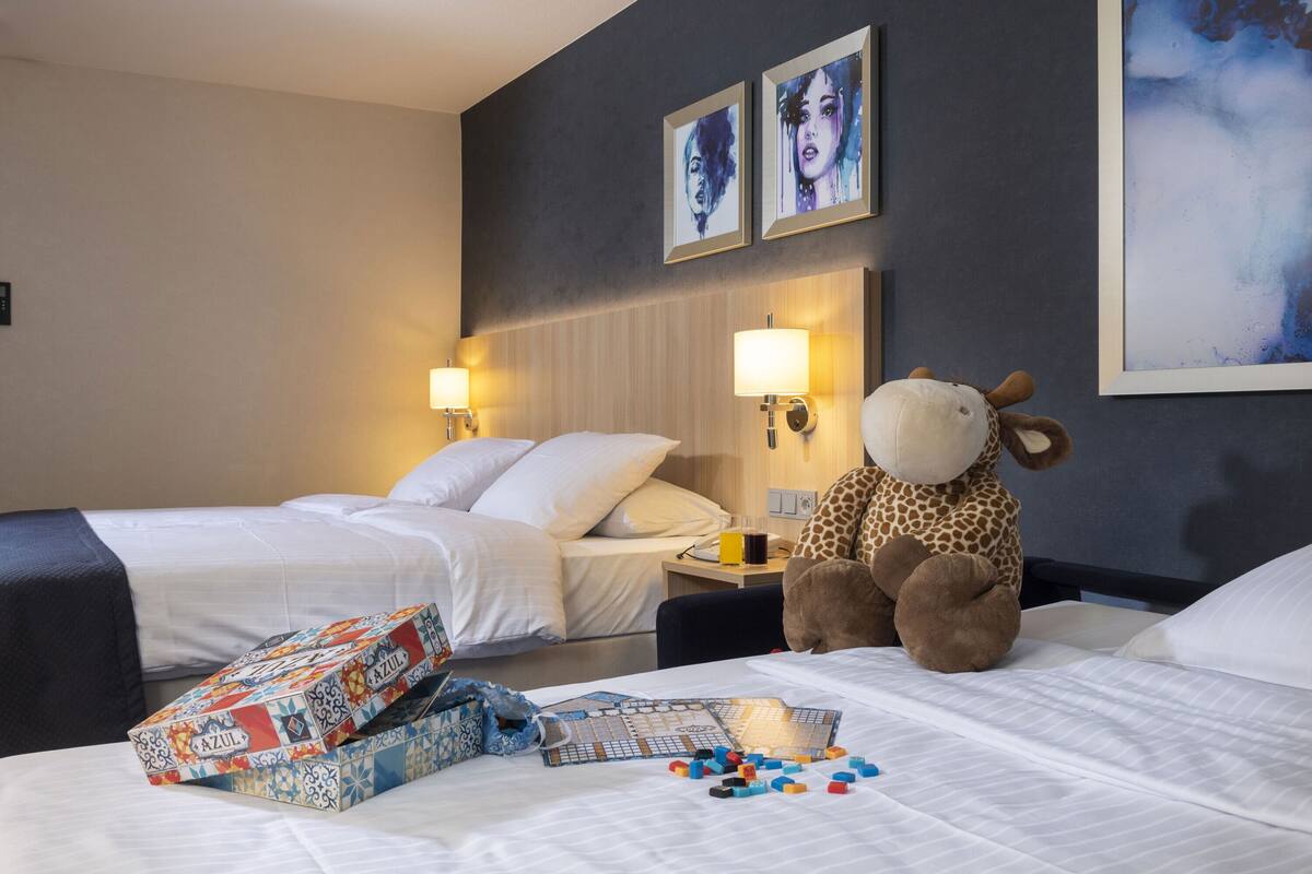 A hotel room with two beds, a large giraffe plush toy, and a board game on the bed.
