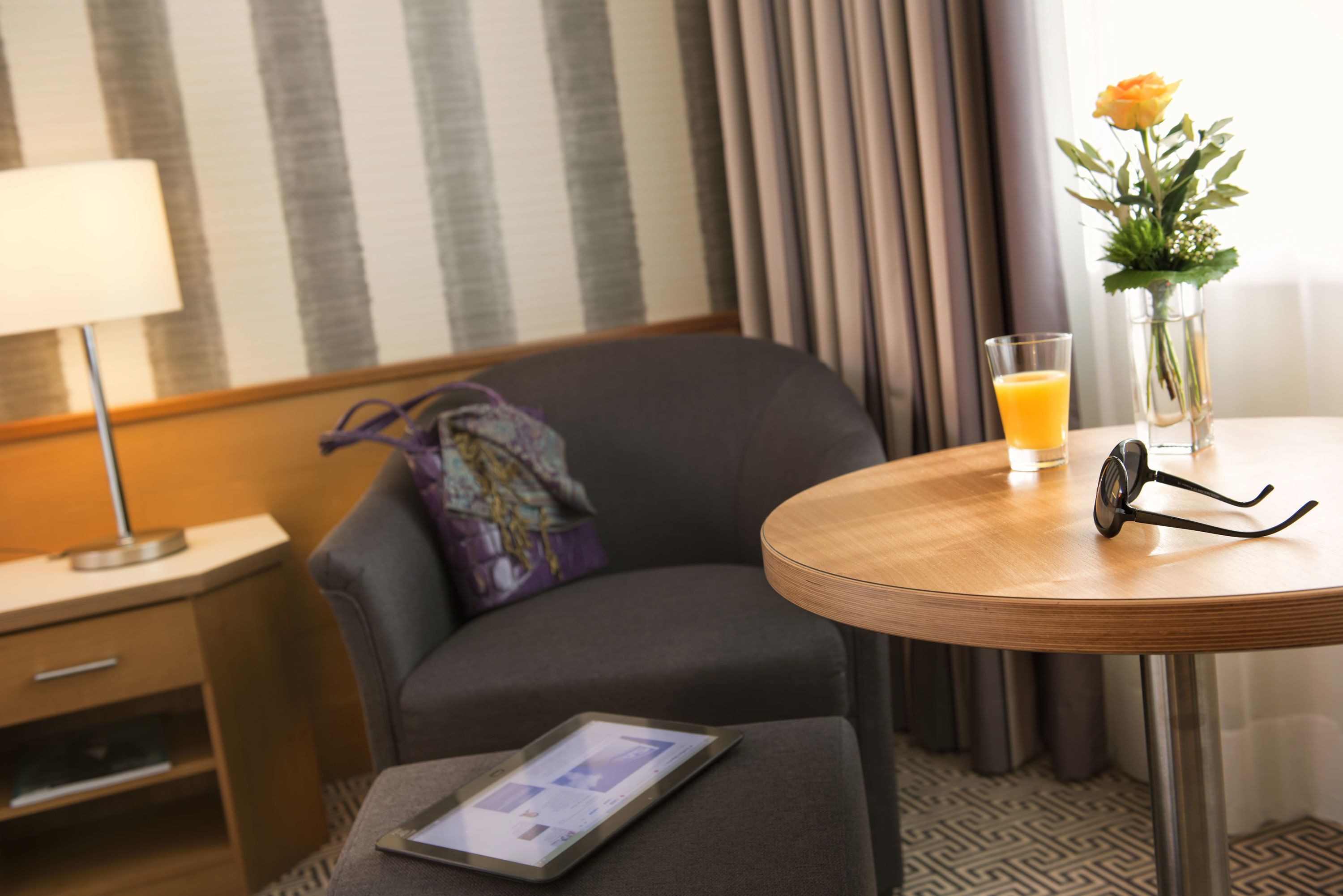 A hotel room with a bed, a bedside table with a lamp, a glass of orange juice, a vase with flowers, and a tablet on a small ottoman.