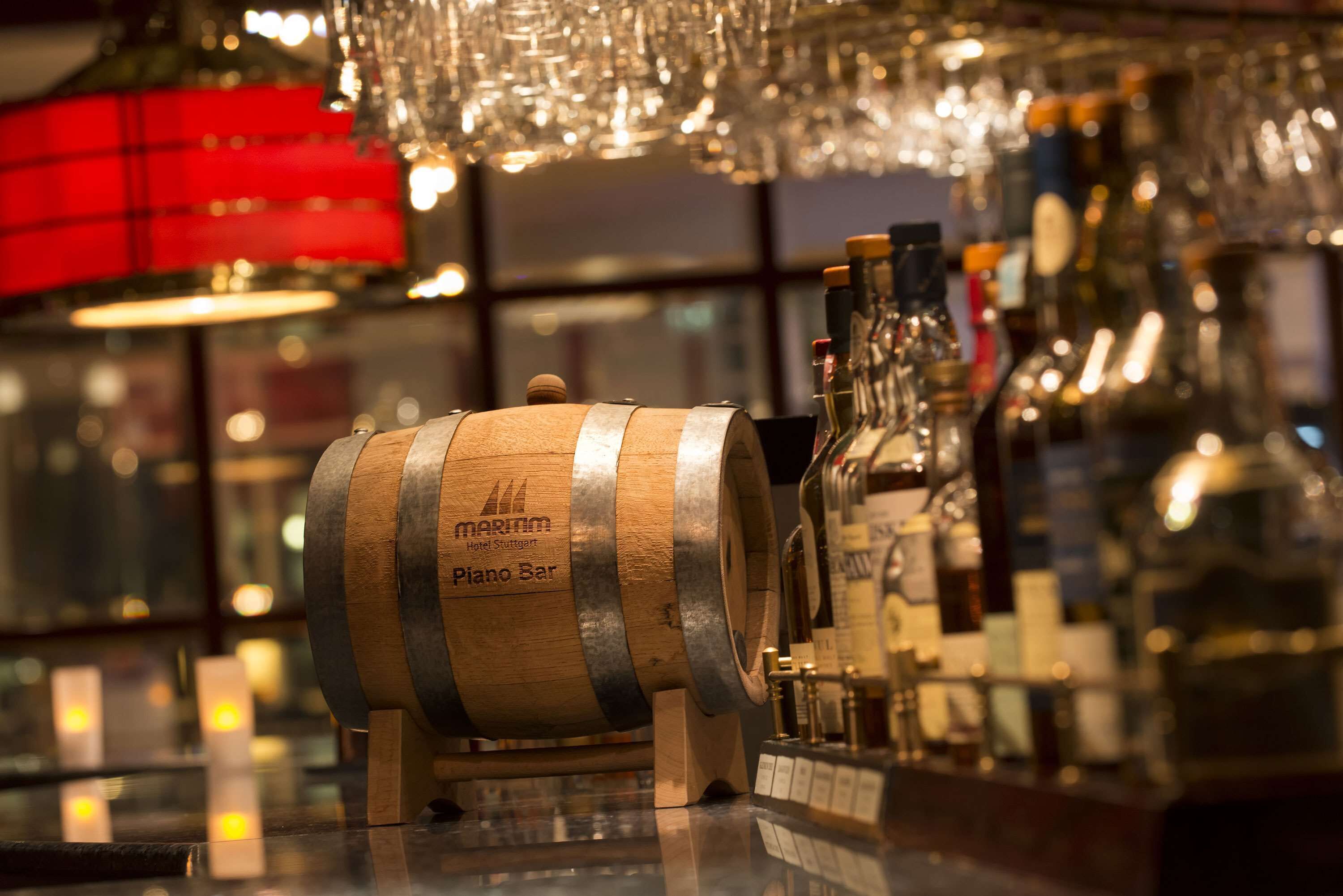 A wooden barrel labeled "Merum Hotel Stuttgart Piano Bar" on a bar counter with various bottles and a chandelier above.