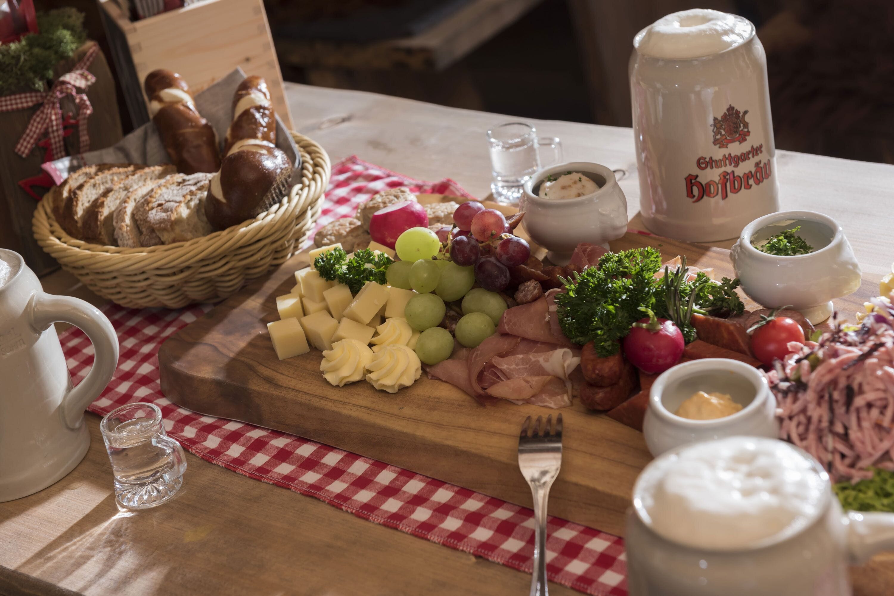 Une planche en bois garnie de différents fromages, de fruits et de charcuteries, accompagnée d’une carafe de bière et de petits ramequins de condiments.