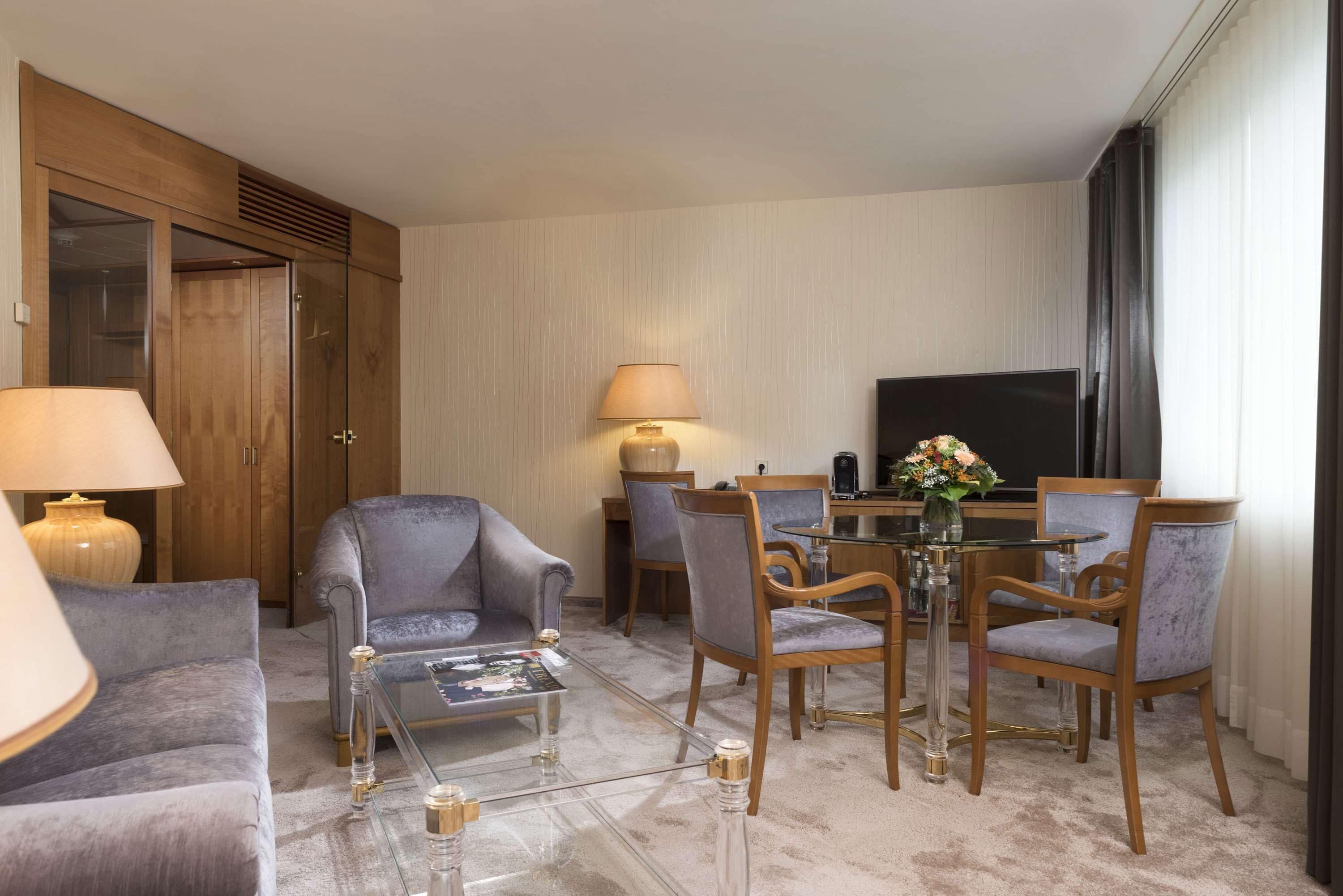 Una habitación de hotel moderna con sofá, sillones, una mesa de centro de vidrio, una mesa de comedor con sillas, un televisor y un armario grande de madera.
