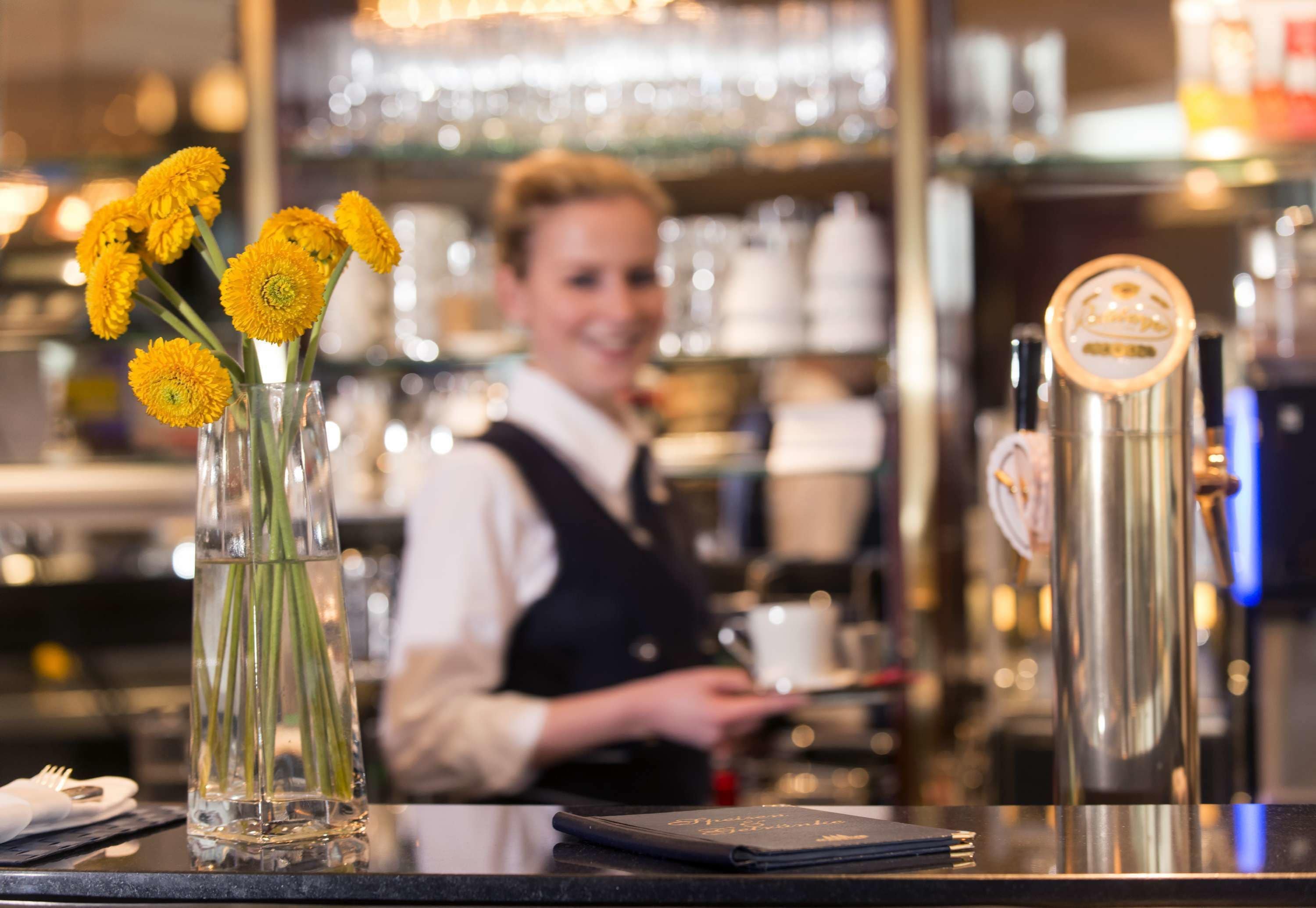 A barista at work, a clear glass vase with yellow flowers, and a stainless steel milk frother on the counter.