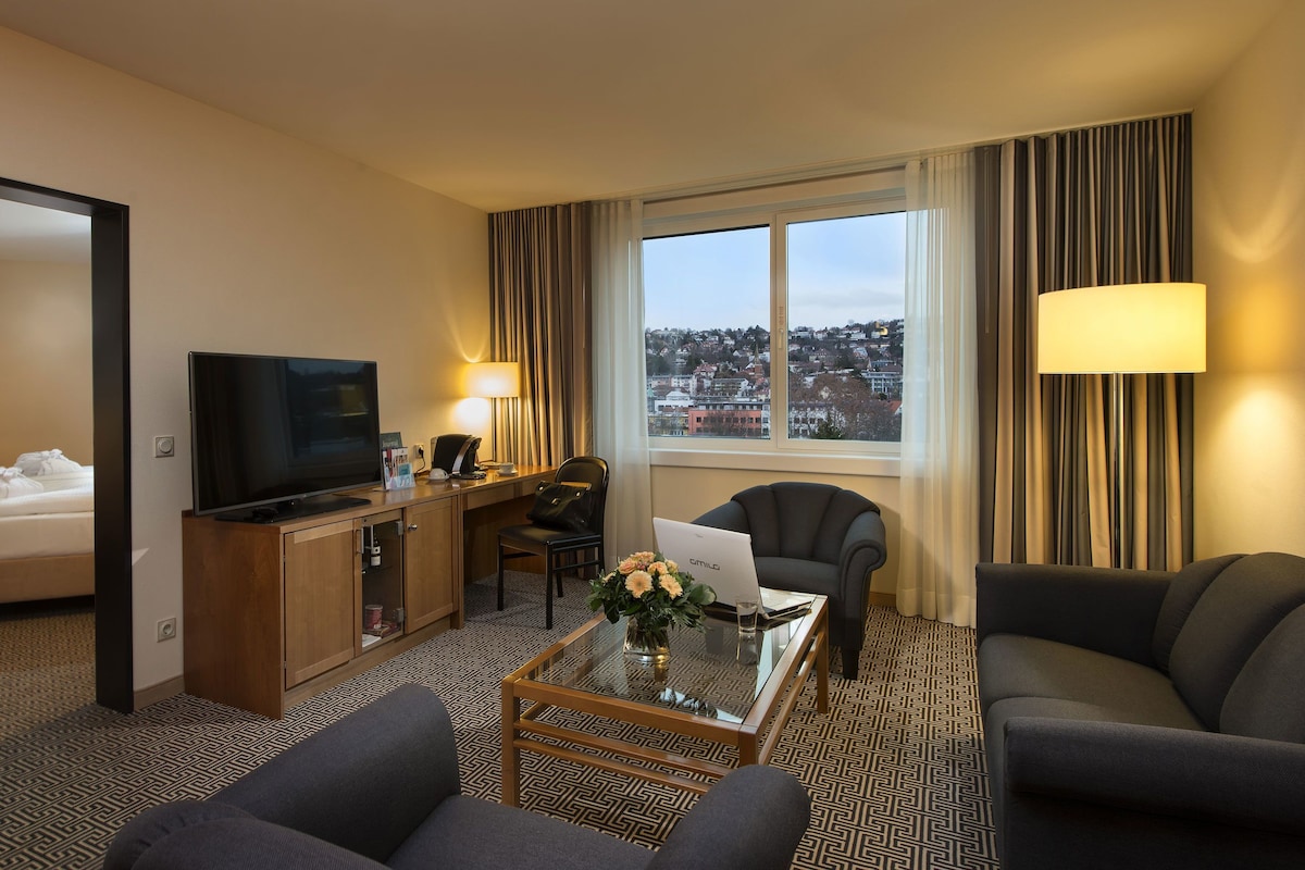 A hotel room with a bed, a desk with a computer, a TV, a coffee table with a vase, and a view of the city.