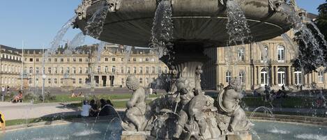 A large ornate fountain with multiple statues, surrounded by a spacious plaza and a historic building in the background.