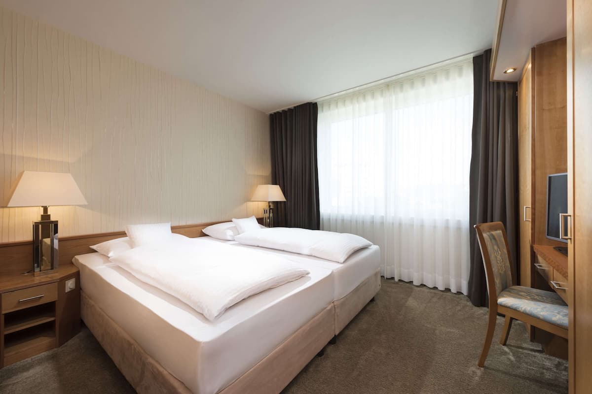 Hotellihuone, jossa on parivuode, kaksi yöpöytää valaisimineen, televisio, tuoli ja suuri ikkuna, jossa on verhot.