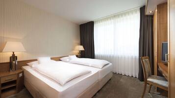 Ein Hotelzimmer mit Doppelbett, zwei Nachttischen mit Lampen, einem Fernseher, einem Sessel und einem großen Fenster mit Vorhängen.