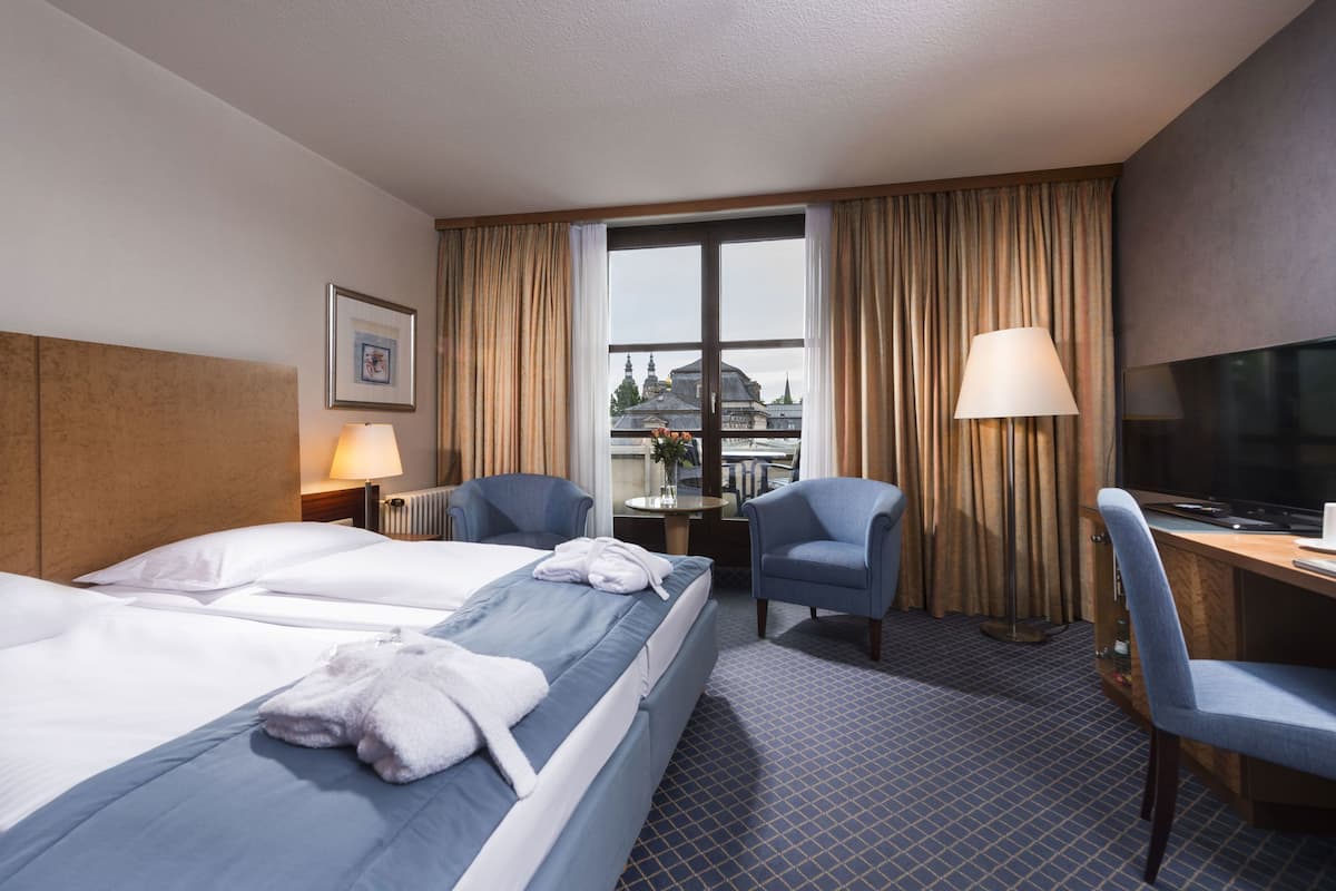 Ein Hotelzimmer mit zwei Betten, einem Schreibtisch mit Fernseher, einem Sessel und einem Fenster mit Vorhängen.