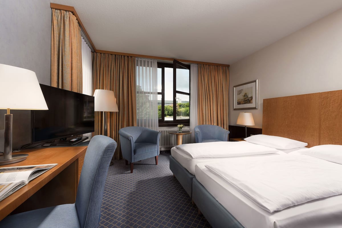 Ein Hotelzimmer mit einem Bett, zwei Sesseln, einem Schreibtisch mit Computer, einem Fernseher und einem Fenster mit Vorhängen.