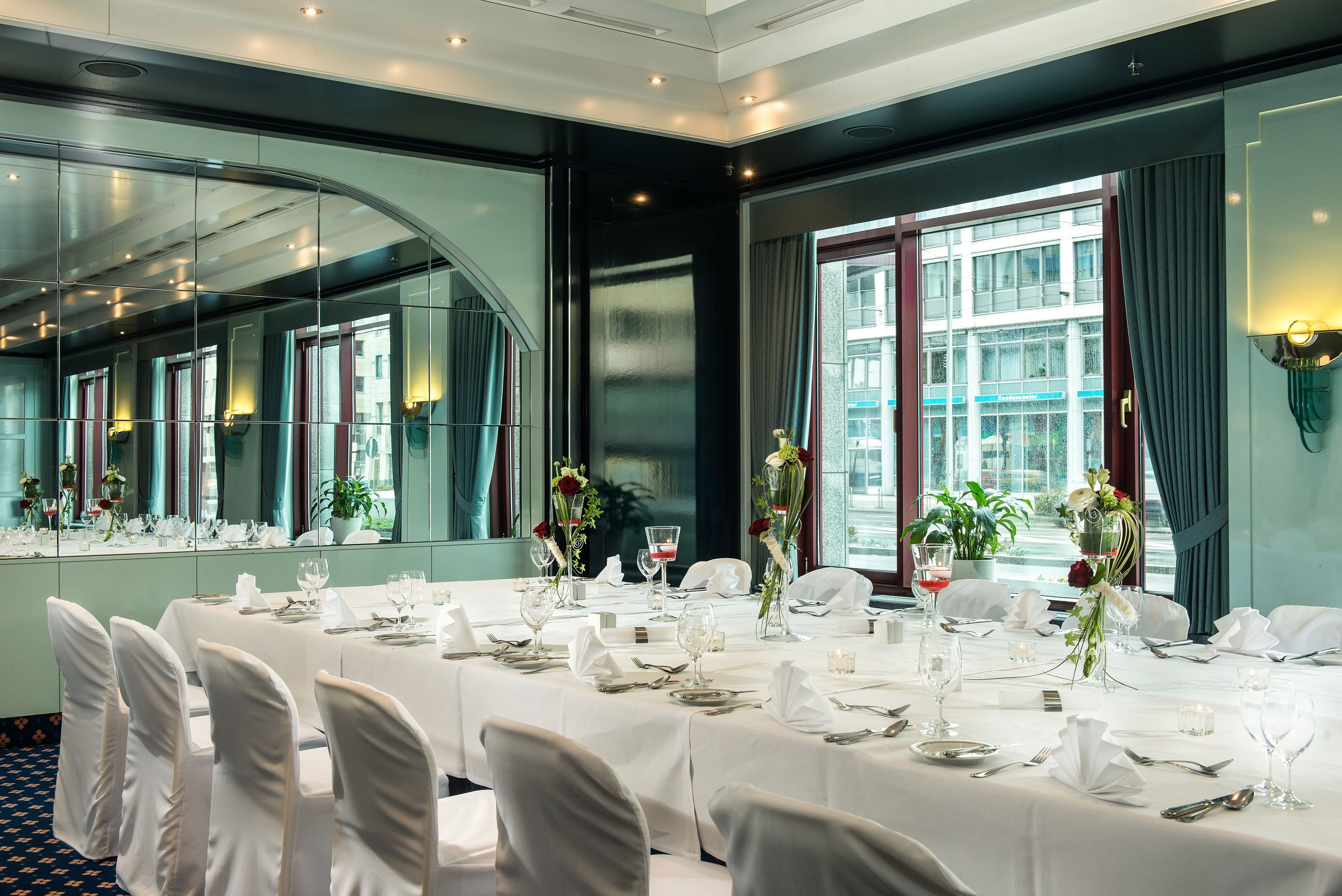 A banquet hall with long tables set for a formal event, large windows, and a mirror reflecting the room.