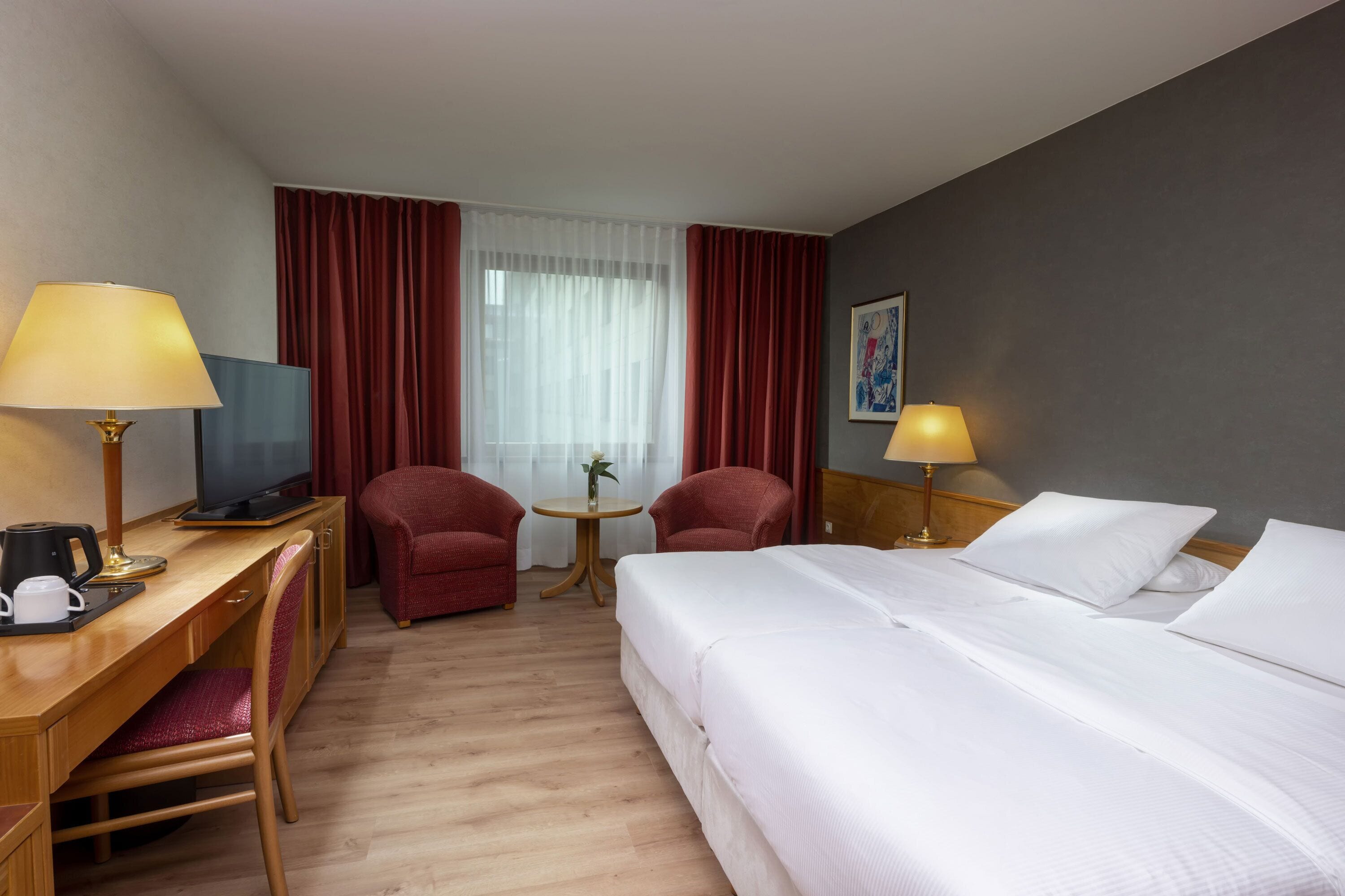 Habitación de hotel con cama, escritorio con computadora, dos sillas, una mesita con un florero y una lámpara.