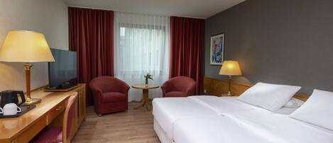 Quarto de hotel com uma cama, uma escrivaninha com computador, duas cadeiras, uma mesinha de centro com um vaso e uma luminária.