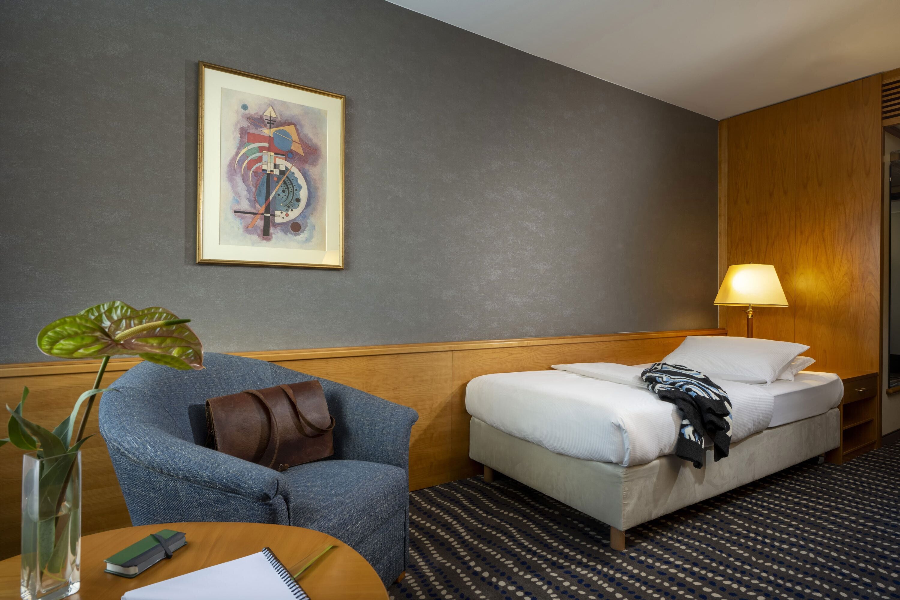 Una habitación de hotel con cama, una silla, un escritorio con lámpara, una obra de arte enmarcada en la pared y una planta.