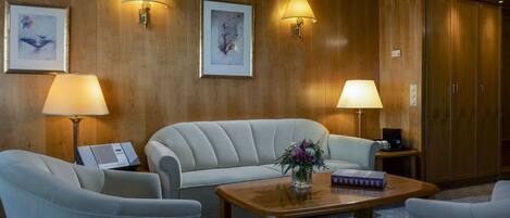 A hotel room with a wooden paneled wall, two wall-mounted lamps, a sofa, a coffee table with a book and flowers, and a wardrobe.