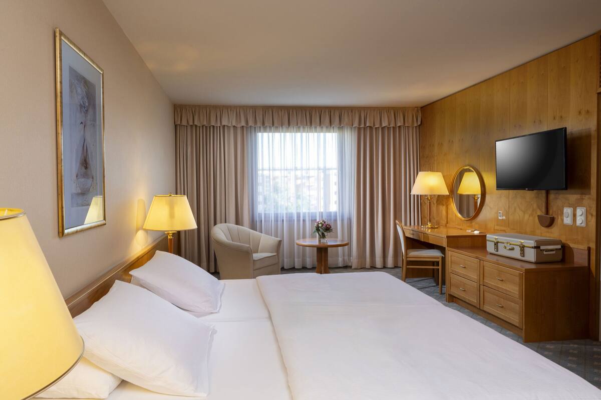 A hotel room with a large bed, two bedside lamps, a desk with a chair, a television, and a window with curtains.