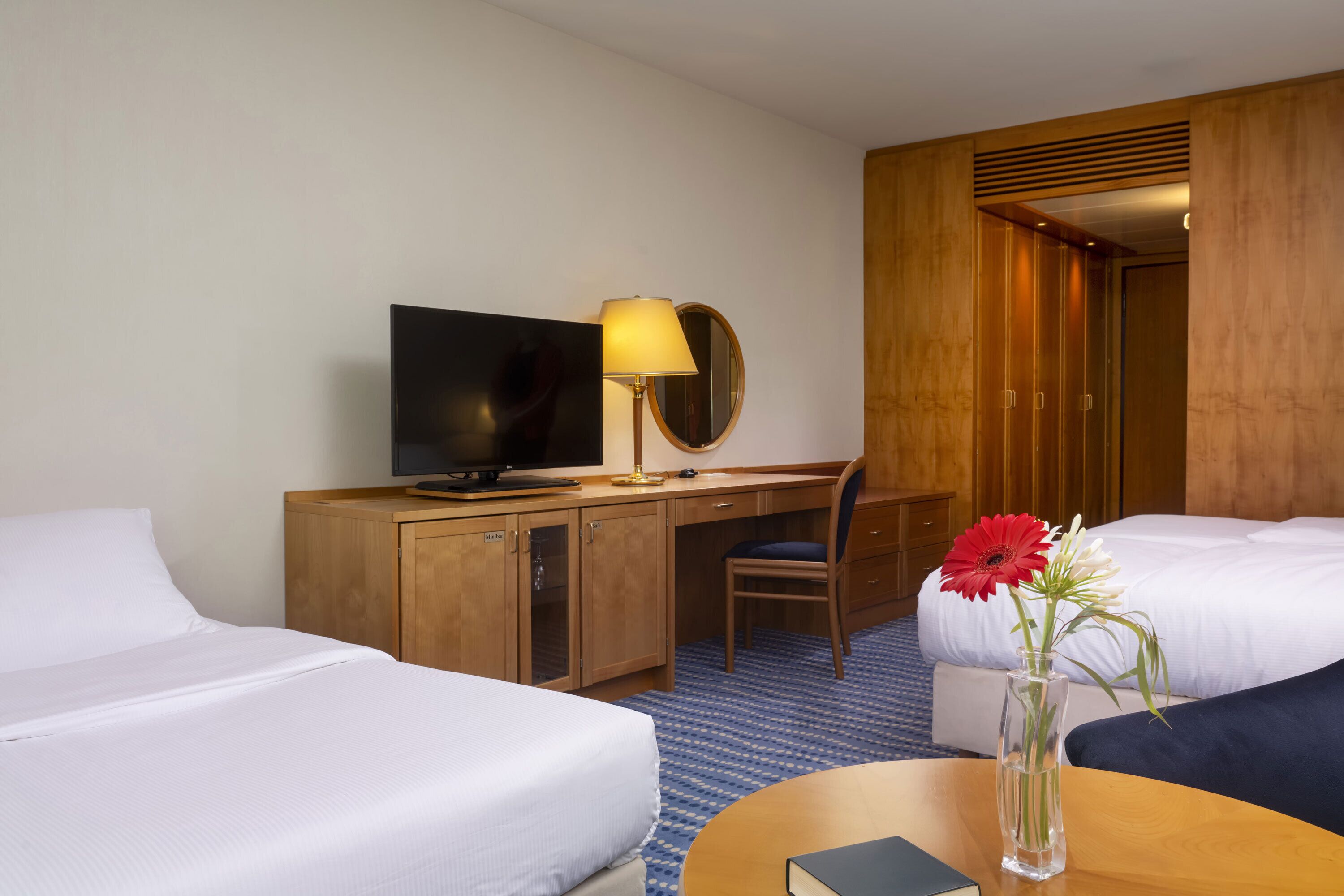A hotel room with two beds, a desk with a TV, a mirror, and a vase with flowers.