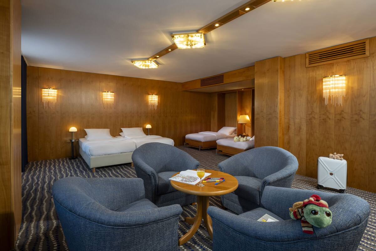 A hotel room with a bed, two armchairs, a small table with a drink and a book, and a suitcase.