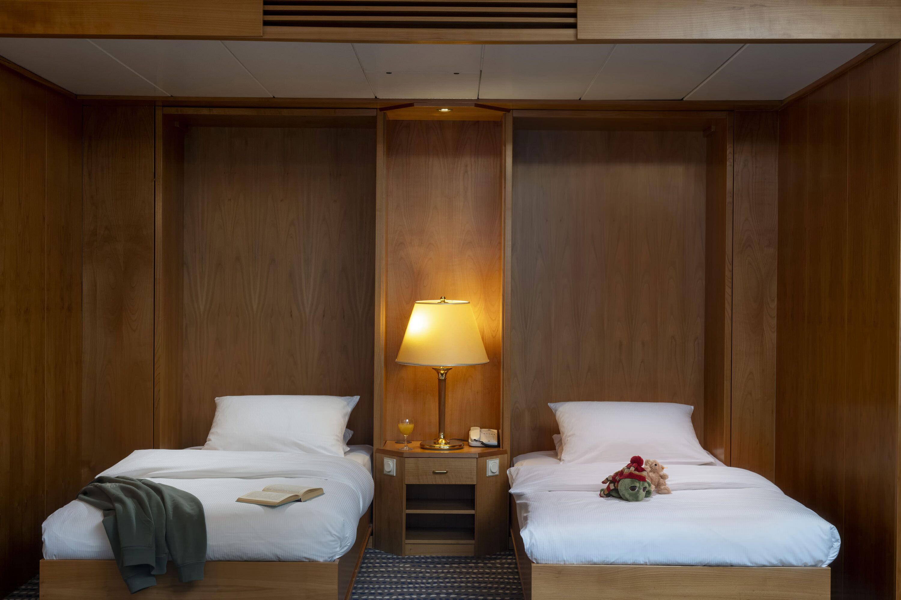 A hotel room with two beds, a nightstand with a lamp, a small table, and a teddy bear on one bed.