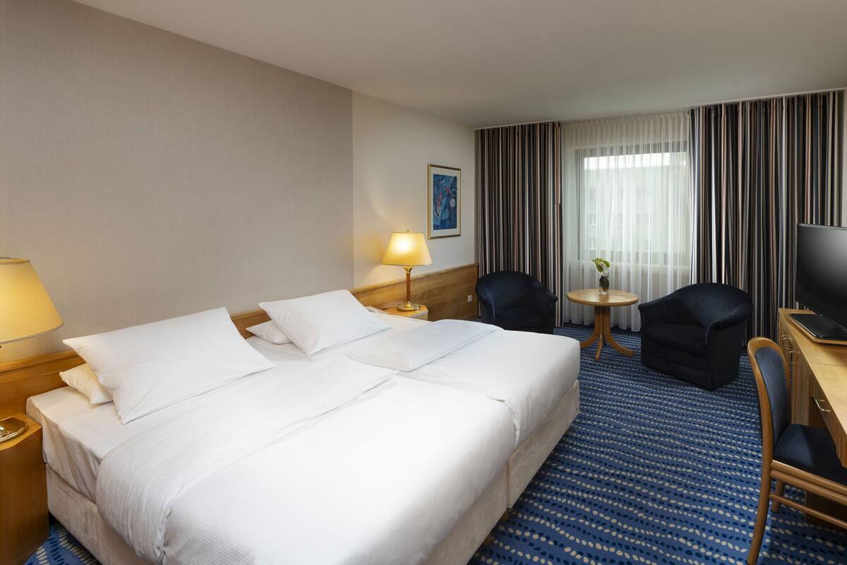 A hotel room with a large bed, two bedside tables with lamps, a desk with a television, a chair, and a small table with a vase.