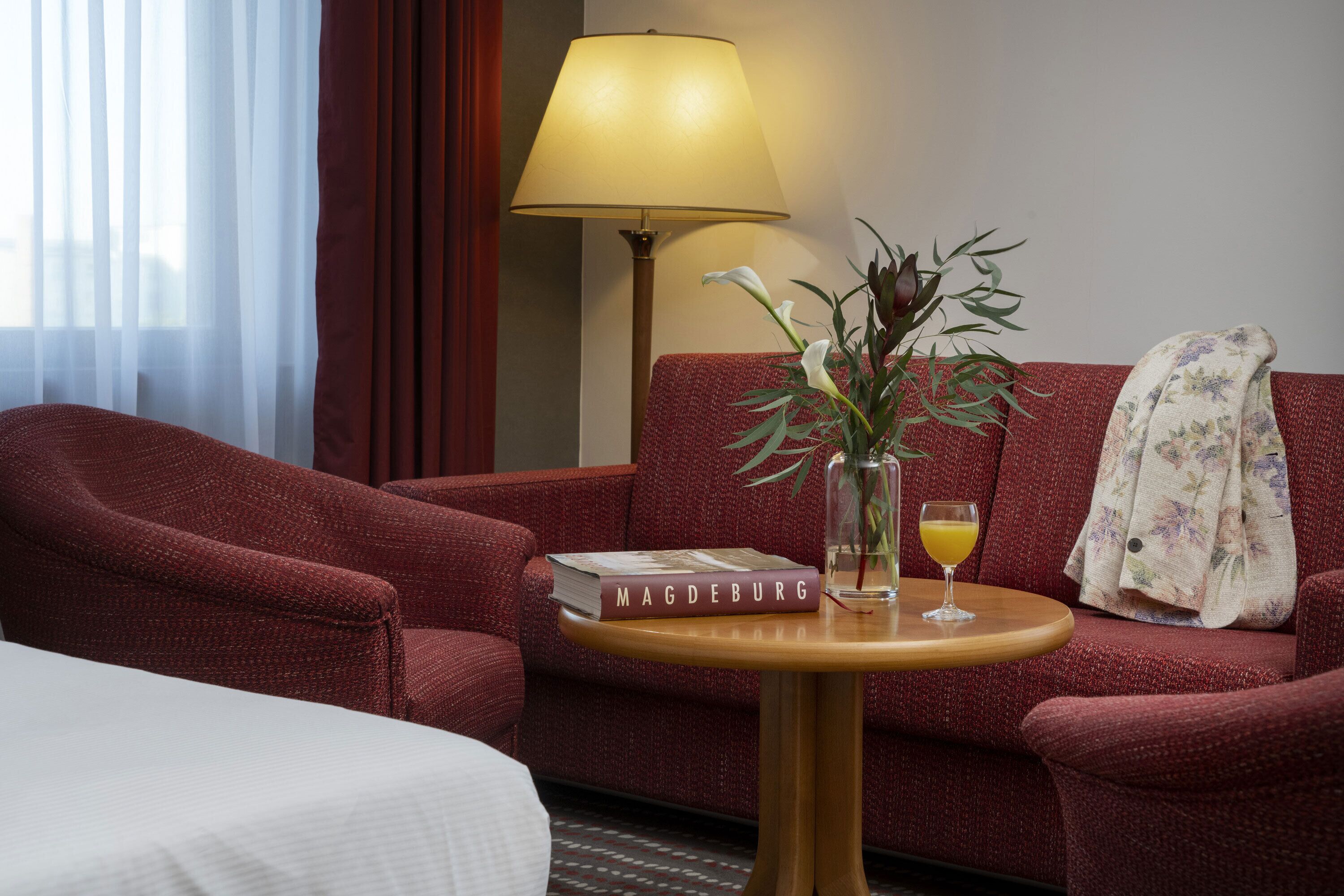 A hotel room with a red sofa, a small round table with a book, a lamp, and a vase with flowers.