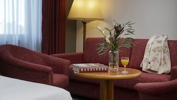 Una habitación de hotel con un sofá rojo, una pequeña mesa redonda con un libro, una lámpara y un jarrón con flores.