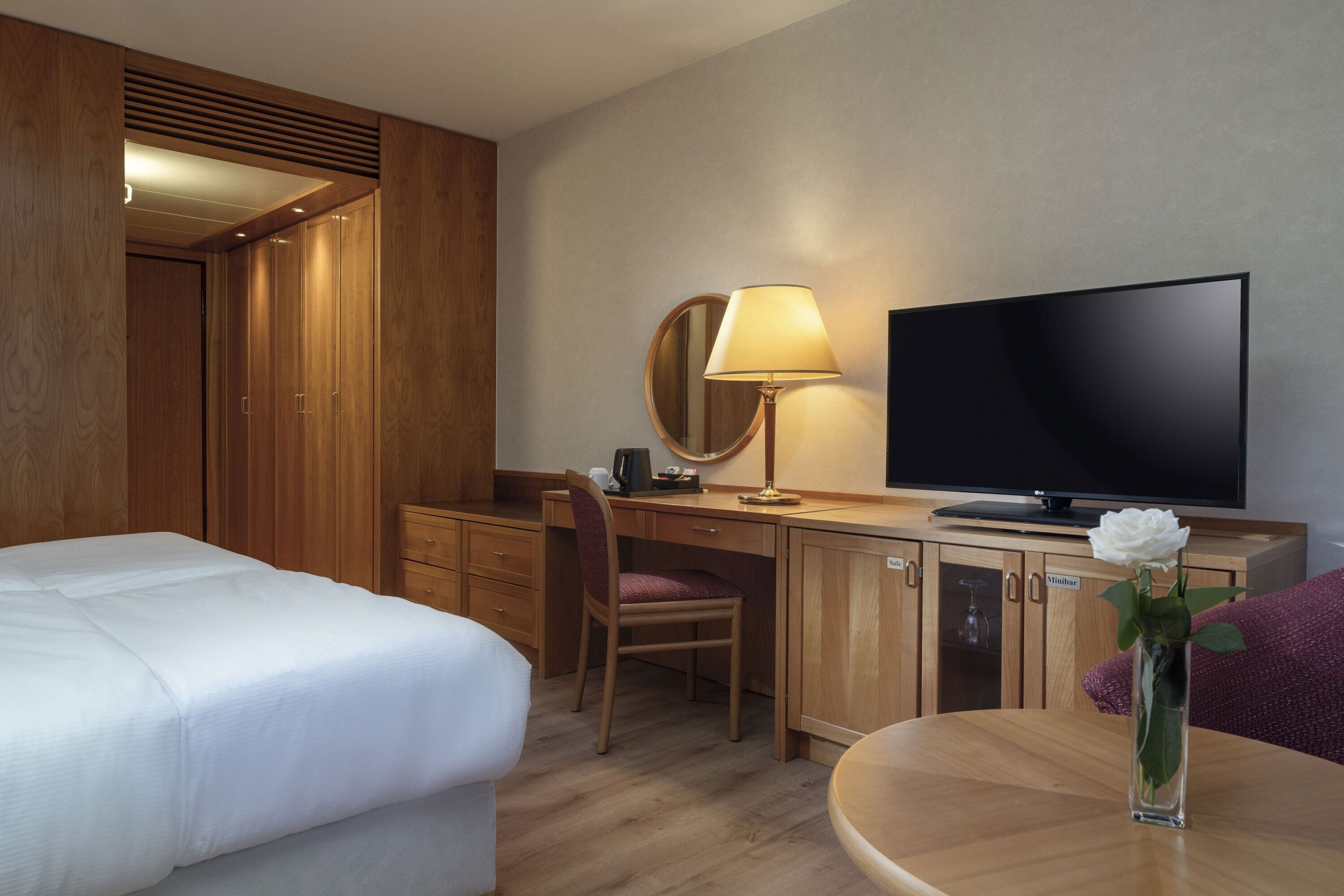 A hotel room with a bed, a desk with a TV, a chair, a lamp, and a vase with a flower.