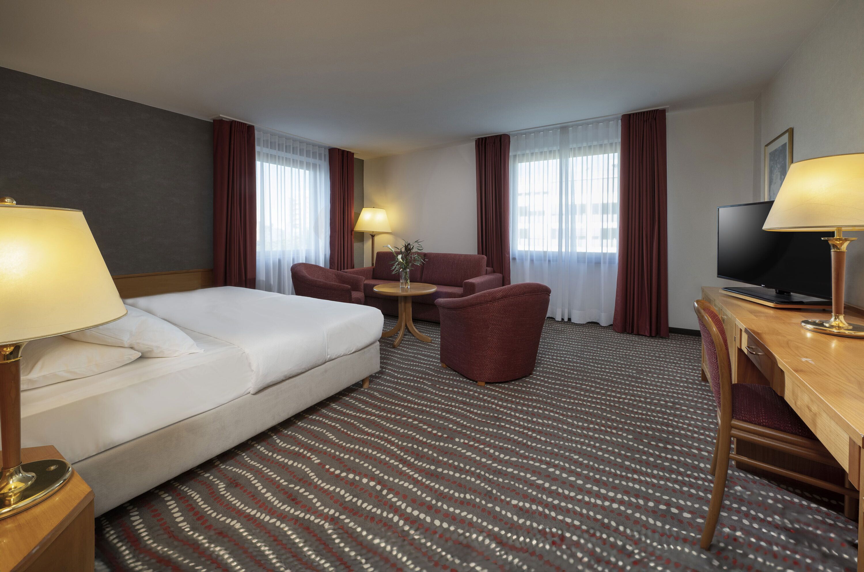A hotel room with a bed, a desk, a chair, a TV, and a small table with a lamp and vase.