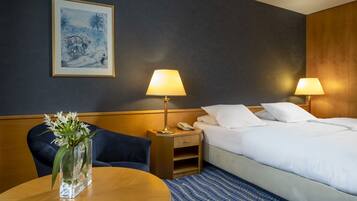 A hotel room with a bed, two bedside lamps, a nightstand, a framed picture on the wall, a vase with flowers on a table, and a blue patterned rug.
