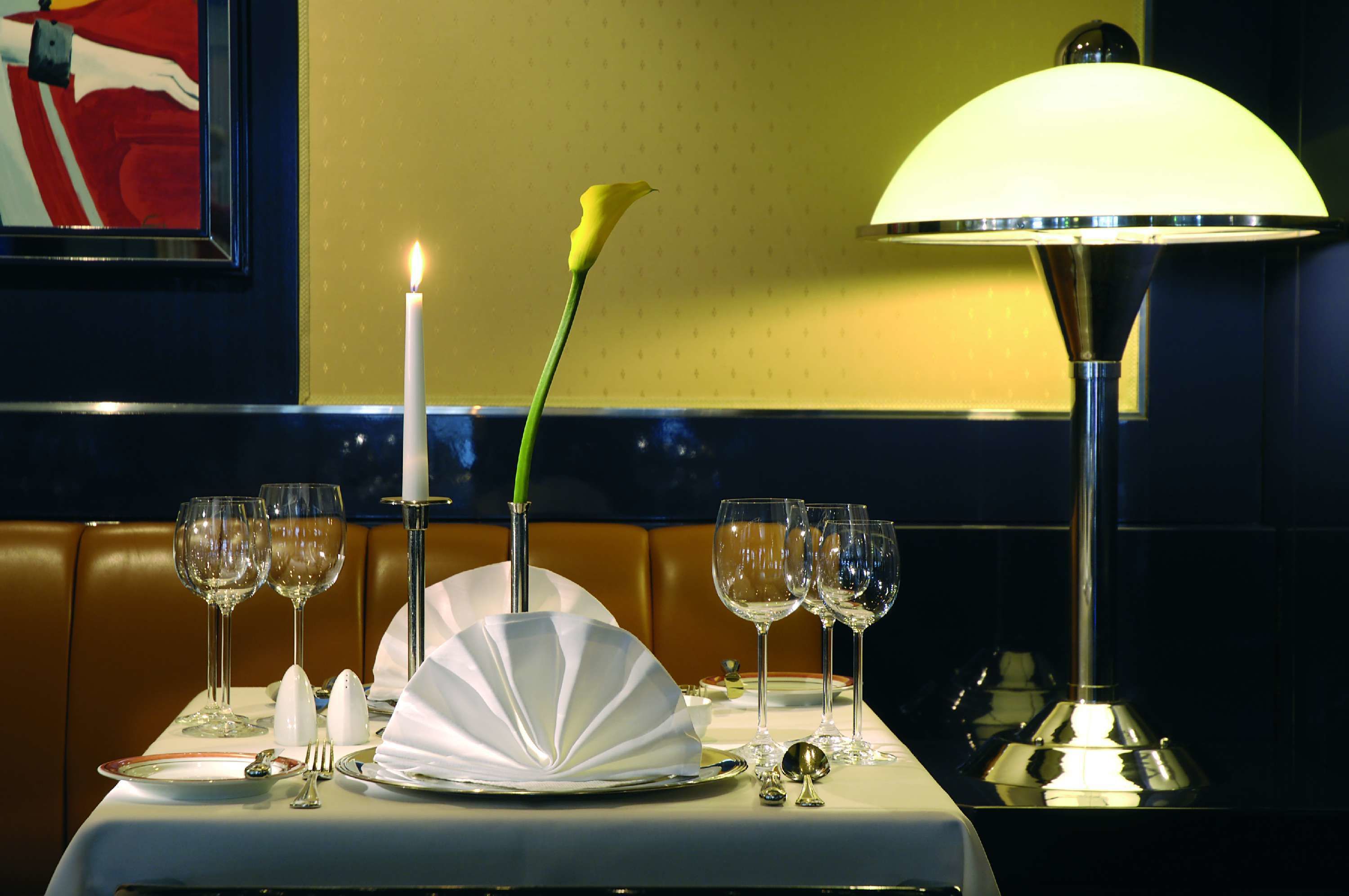 Una mesa de comedor dispuesta con platos blancos, cubiertos y copas de vino, una vela encendida y una flor amarilla.