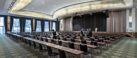 A large conference room with rows of tables and chairs, a stage with curtains, and a patterned carpet.