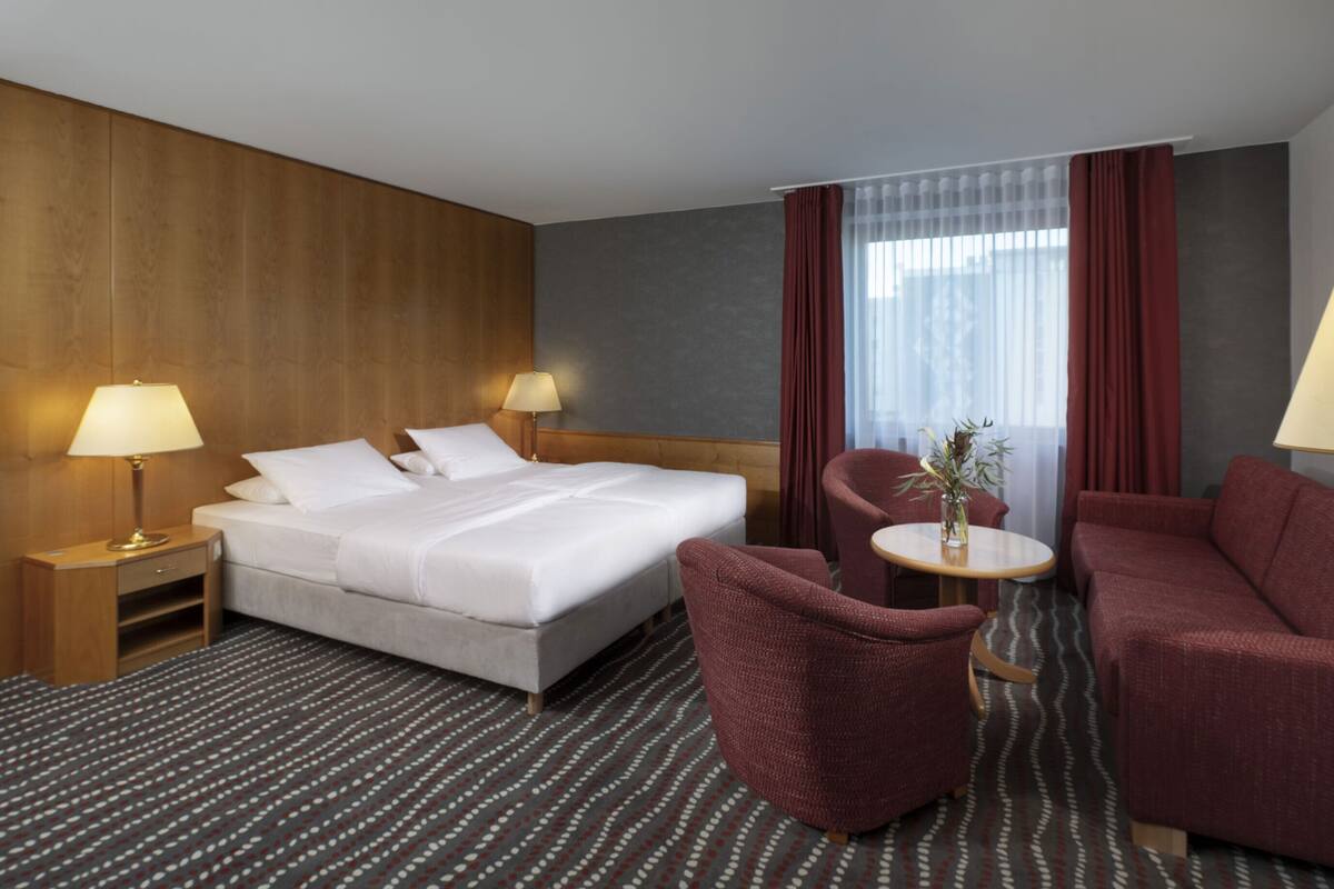 A hotel room with a bed, two bedside tables with lamps, a small round table with a vase, and two armchairs.