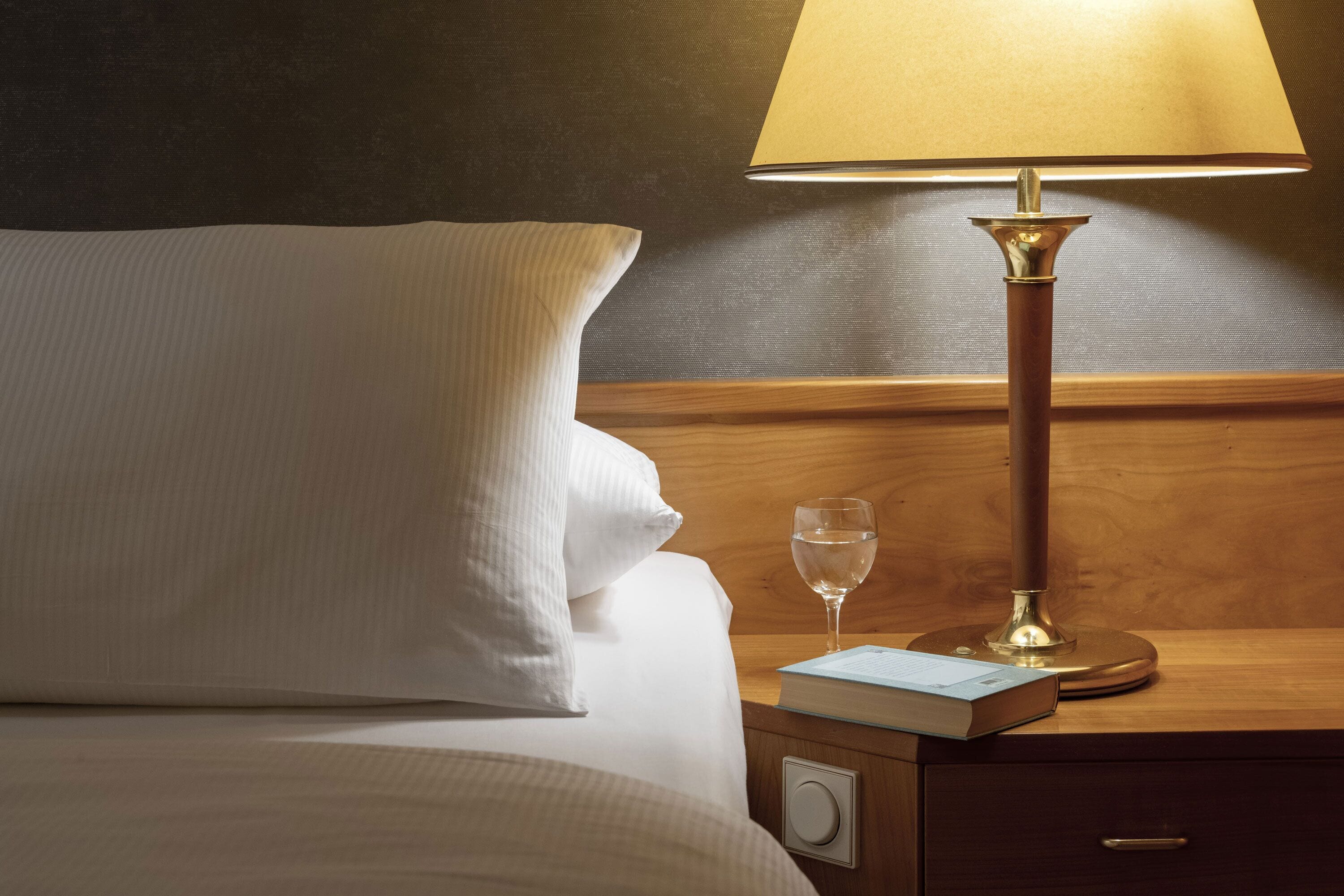A neatly made bed with a pillow, a bedside table with a lamp, a glass of water, and a book.