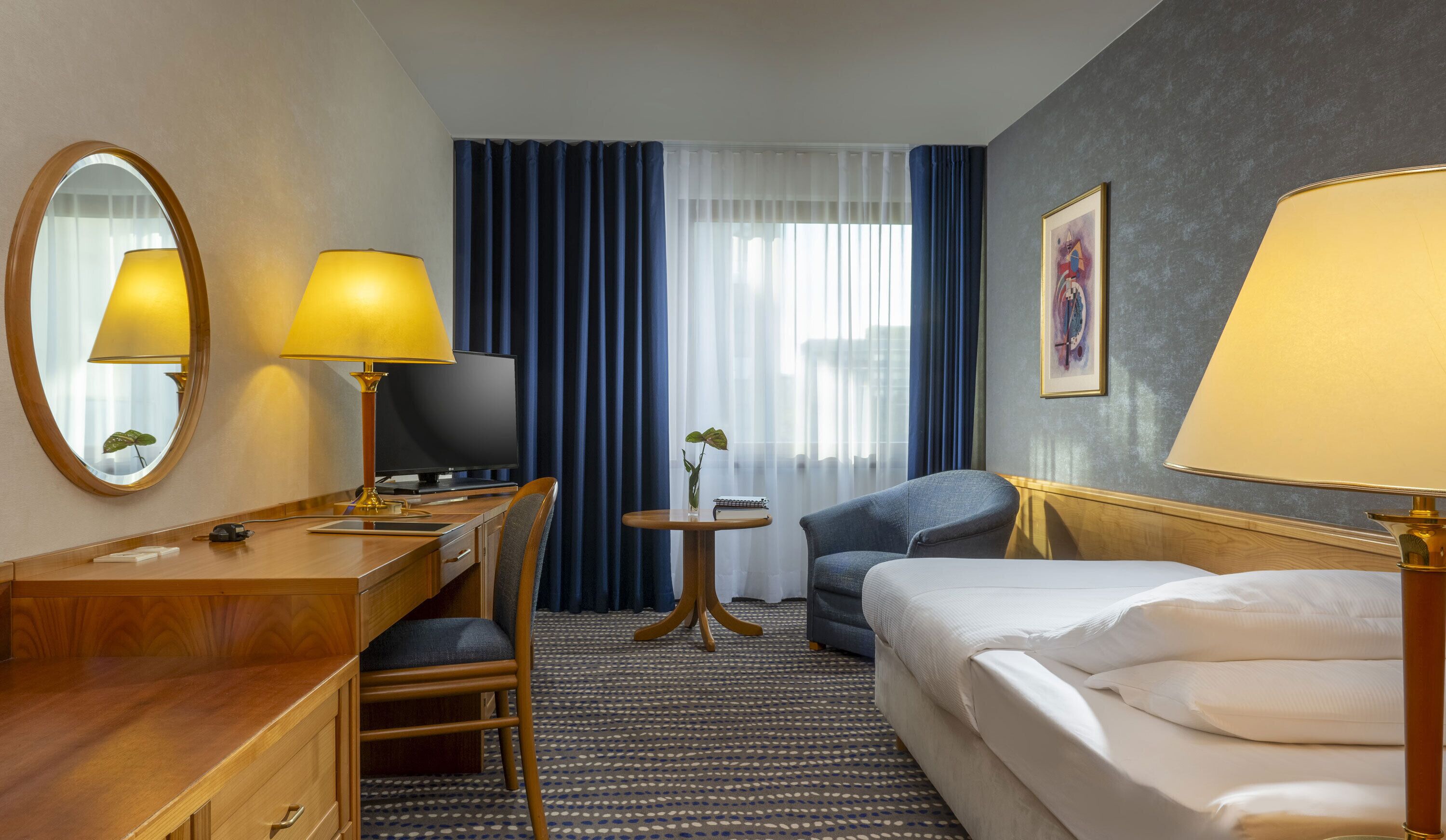 A hotel room with a bed, a desk with a lamp, a chair, a TV, a small table with a plant, and a painting on the wall.