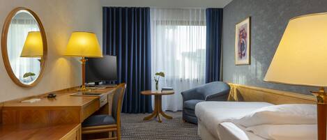 A hotel room with a bed, a desk with a lamp, a chair, a TV, a small table with a plant, and a painting on the wall.